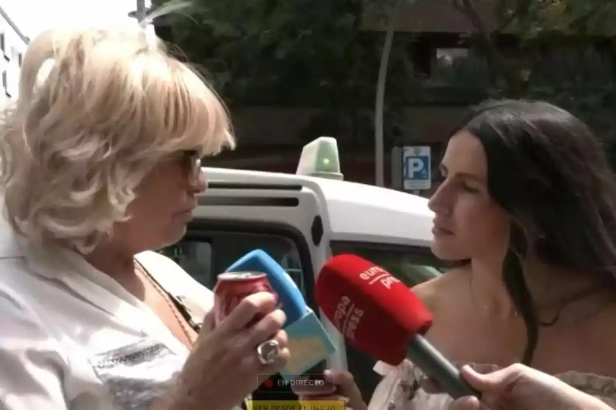 Dos mujeres conversan en la calle mientras Bárbara Rey sostiene una lata de refresco y la otra varios micrófonos de prensa como La Familia de la Tele frente a un taxi blanco. Dos mujeres conversan en la calle mientras Bárbara Rey sostiene una lata de refresco y la otra varios micrófonos de prensa como La Familia de la Tele frente a un taxi blanco.