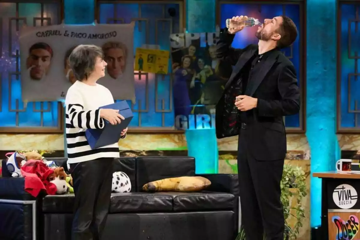 Belén Benito con suéter de rayas sostiene una caja azul mientras David Broncano bebe de una botella en La Revuelta con decoraciones coloridas y un sofá lleno de peluches. Belén Benito con suéter de rayas sostiene una caja azul mientras David Broncano bebe de una botella en La Revuelta con decoraciones coloridas y un sofá lleno de peluches.