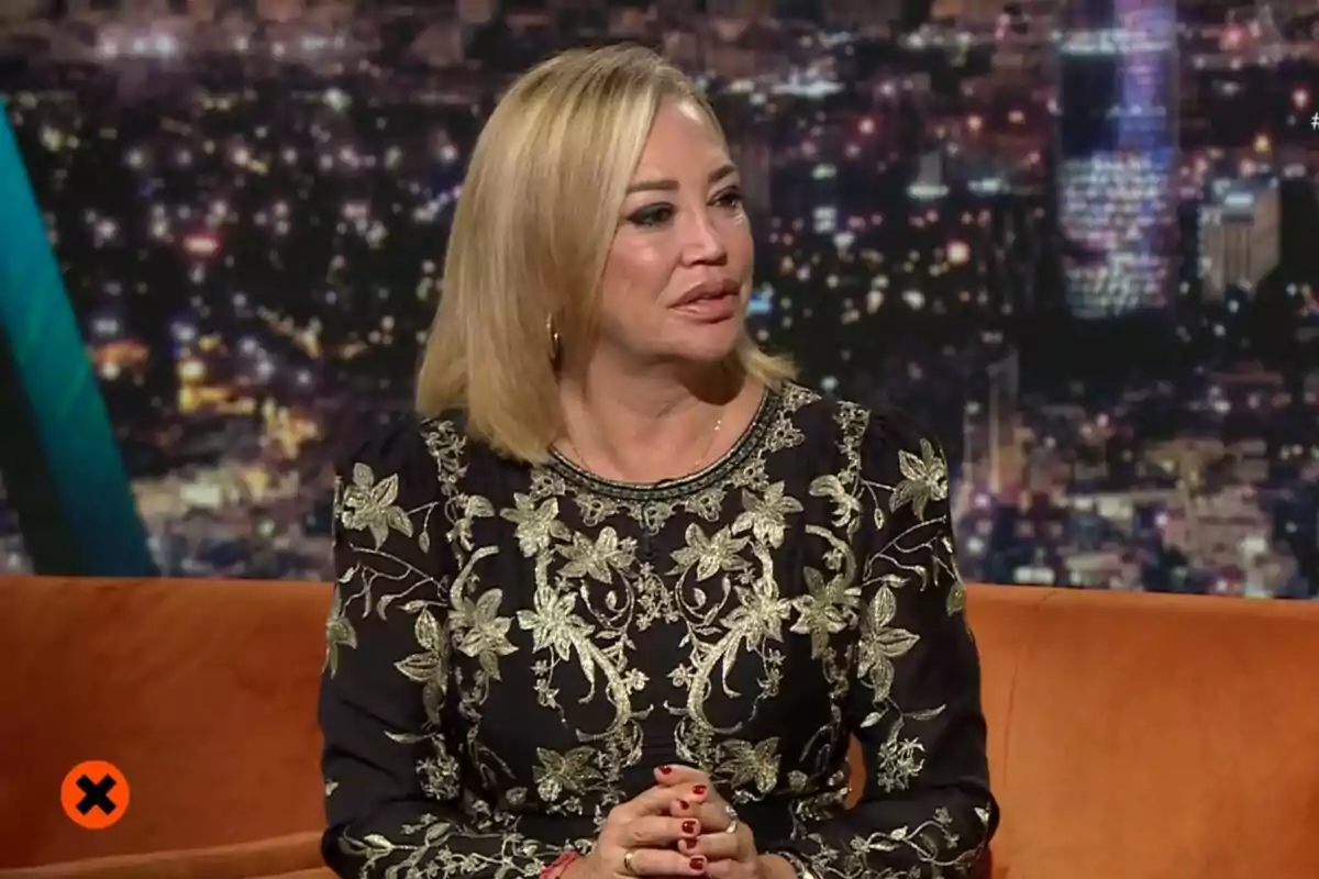 Belén Esteban amb cabell ros i vestit negre amb detalls daurats està asseguda en un sofà taronja amb un fons de llums de ciutat a Col·lapse. Belén Esteban amb cabell ros i vestit negre amb detalls daurats està asseguda en un sofà taronja amb un fons de llums de ciutat a Col·lapse.