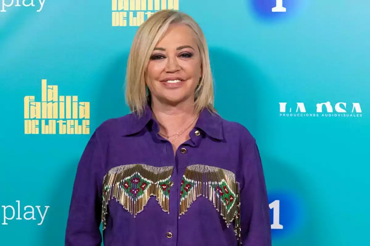 Belén Esteban amb cabell ros i camisa morada decorada, posant davant d'un fons blau amb logotips de 