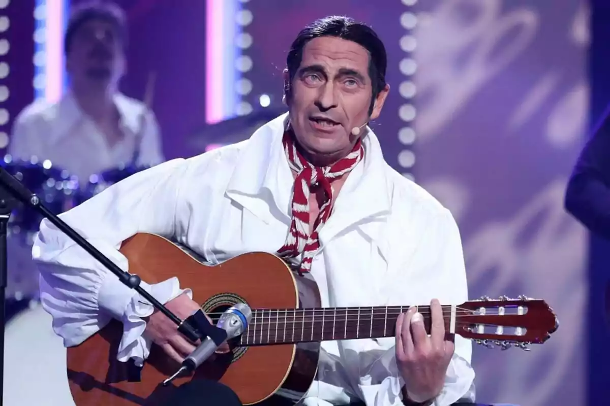 Bertín Osborne con camisa blanca y pañuelo rojo toca la guitarra en un escenario iluminado en Tu Cara Me Suena. Bertín Osborne con camisa blanca y pañuelo rojo toca la guitarra en un escenario iluminado en Tu Cara Me Suena.