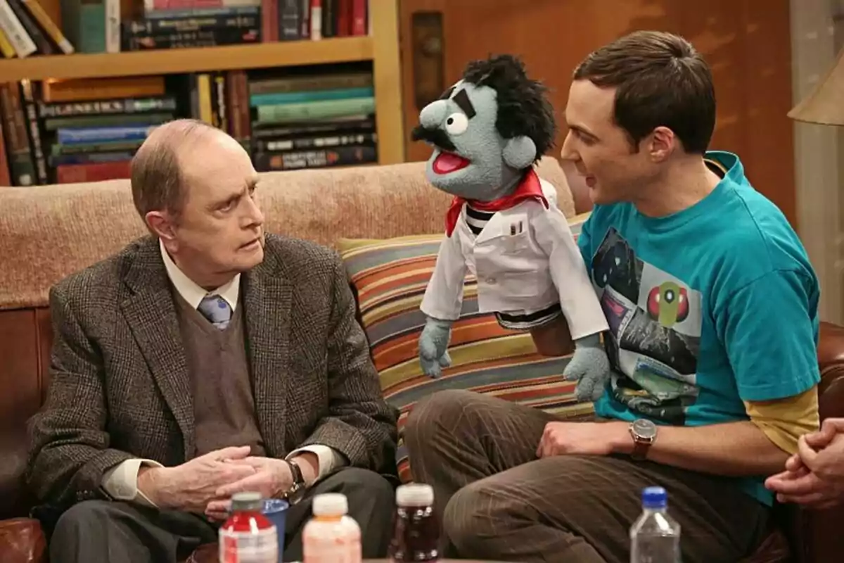 Fotografía de Bob Newhart con traje y corbata está sentado en un sofá junto a Jim Parsons que sostiene un títere de mano vestido con una bata de laboratorio, con una estantería llena de libros al fondo en The Big Bang Theory Fotografía de Bob Newhart con traje y corbata está sentado en un sofá junto a Jim Parsons que sostiene un títere de mano vestido con una bata de laboratorio, con una estantería llena de libros al fondo en The Big Bang Theory