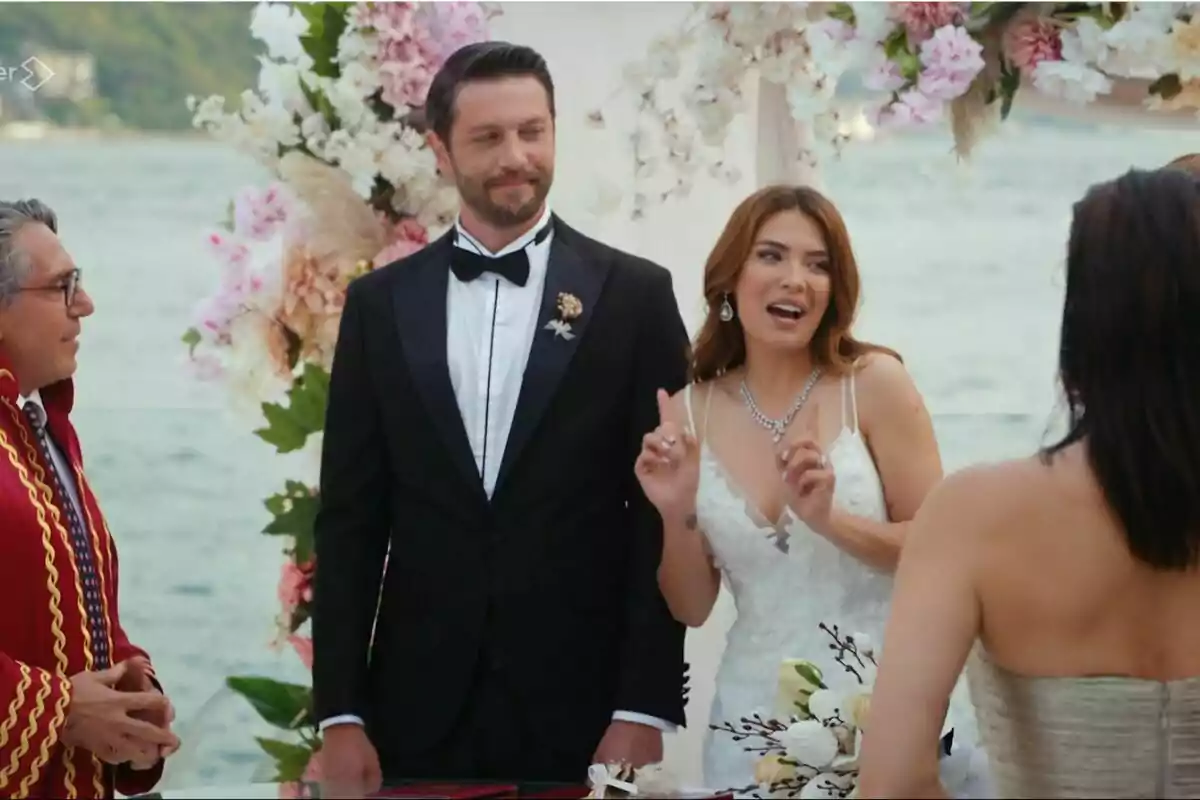 Çaner y Kumru vestidos de novios está en una ceremonia al aire libre junto a un oficiante y otra persona, con un fondo de flores y el mar en Pecado Original Çaner y Kumru vestidos de novios está en una ceremonia al aire libre junto a un oficiante y otra persona, con un fondo de flores y el mar en Pecado Original