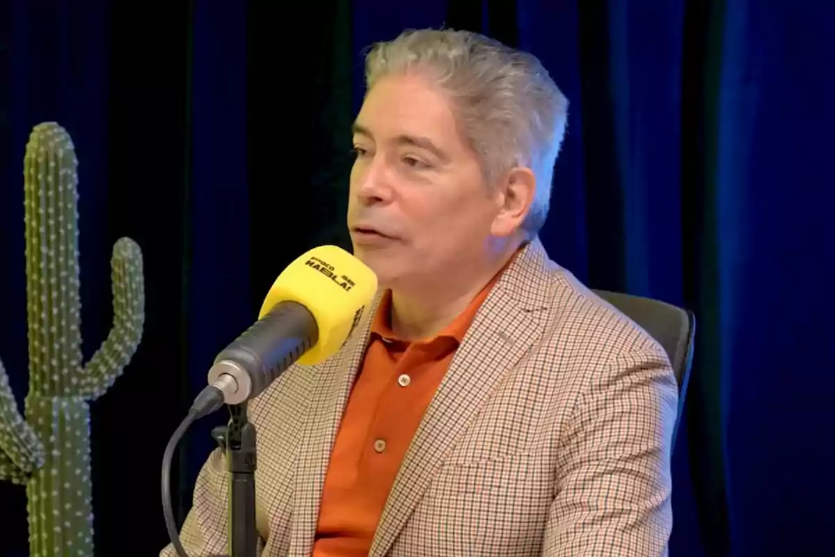 Boris Izaguirre de cabello canoso y chaqueta a cuadros habla frente a un micrófono amarillo de Poco se Habla! en un estudio con fondo azul y un cactus a su lado. Boris Izaguirre de cabello canoso y chaqueta a cuadros habla frente a un micrófono amarillo de Poco se Habla! en un estudio con fondo azul y un cactus a su lado.