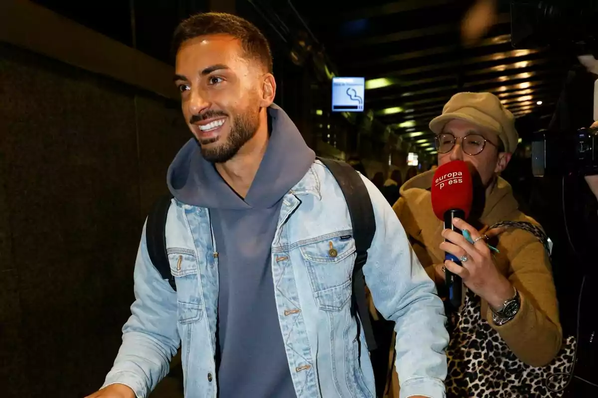 Borja González sonriente con chaqueta de mezclilla y mochila es entrevistado por un reportero con micrófono rojo en el aeropuerto camino a Supervivientes. Borja González sonriente con chaqueta de mezclilla y mochila es entrevistado por un reportero con micrófono rojo en el aeropuerto camino a Supervivientes.