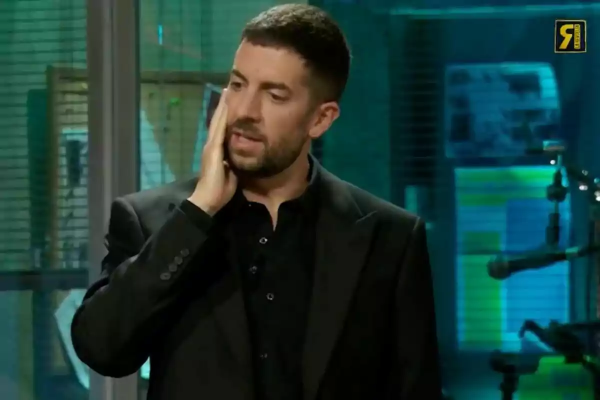 David Broncano con barba y cabello corto, vestido con un traje negro, se toca la mejilla mientras está de pie en un entorno interior con iluminación azul y verde en 'La Revuelta'.