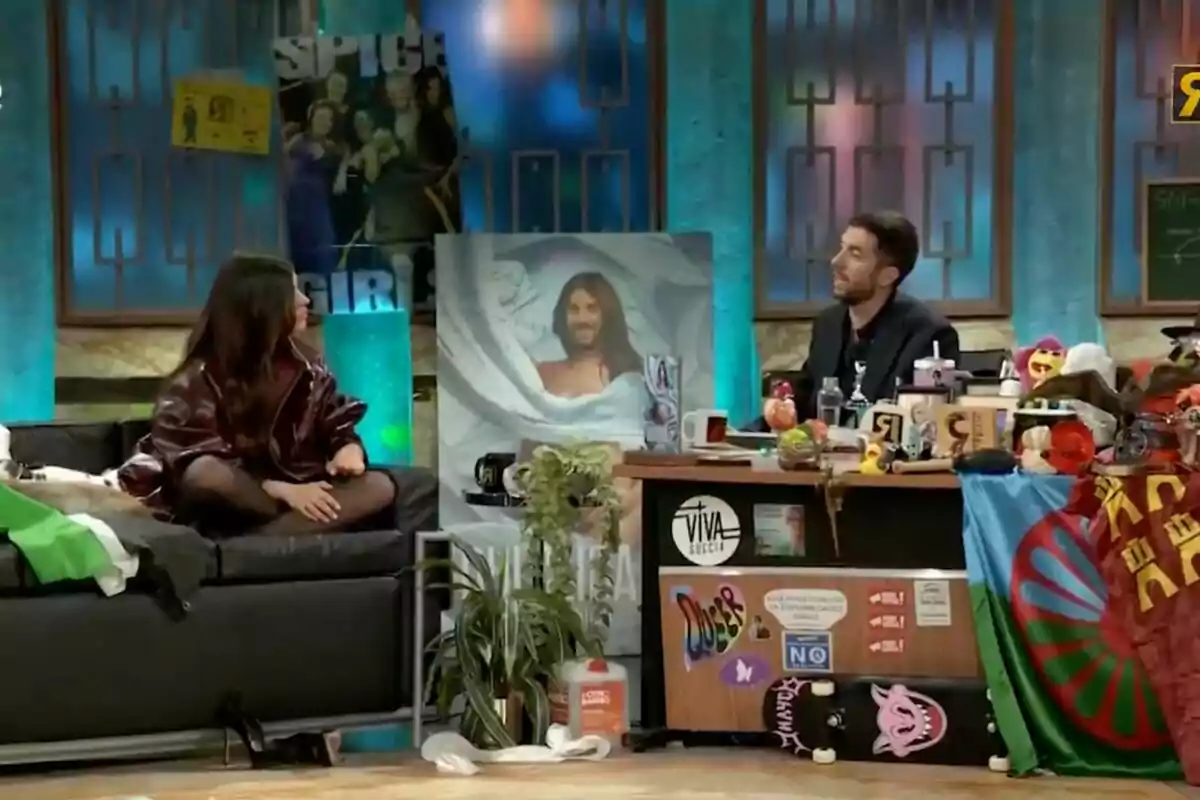 En un plató de televisió, Dulceida està asseguda en un sofà davant de David Broncano, que és darrere d'un escriptori ple d'objectes variats, incloent-hi tasses, joguines i una bandera; a la paret hi ha un pòster de les Spice Girls i una imatge d'una persona embolicada en un llençol a 'La Revuelta'. En un plató de televisió, Dulceida està asseguda en un sofà davant de David Broncano, que és darrere d'un escriptori ple d'objectes variats, incloent-hi tasses, joguines i una bandera; a la paret hi ha un pòster de les Spice Girls i una imatge d'una persona embolicada en un llençol a 'La Revuelta'.
