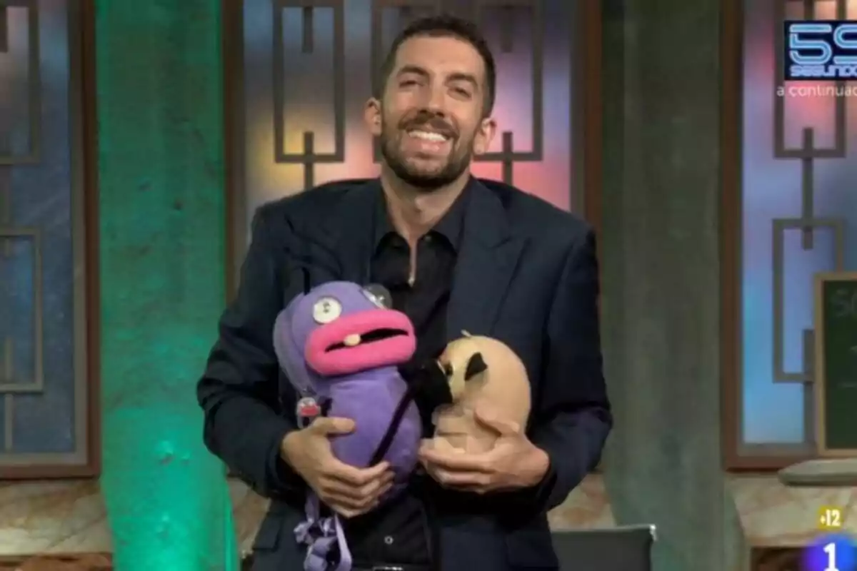 David Broncano sonriente en La Revuelta sostiene dos marionetas, una morada con labios rosados que es una hormiga de EL Hormiguero y otra beige con orejas negras, en un set de televisión con fondo verde y elementos decorativos geométricos. David Broncano sonriente en La Revuelta sostiene dos marionetas, una morada con labios rosados que es una hormiga de EL Hormiguero y otra beige con orejas negras, en un set de televisión con fondo verde y elementos decorativos geométricos.