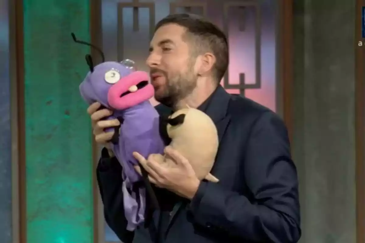 David Broncano con barba y traje en La Revuelta sostiene y besa a una marioneta morada de una hormiga de El Hormiguero con grandes labios rosados y ojos saltones. David Broncano con barba y traje en La Revuelta sostiene y besa a una marioneta morada de una hormiga de El Hormiguero con grandes labios rosados y ojos saltones.