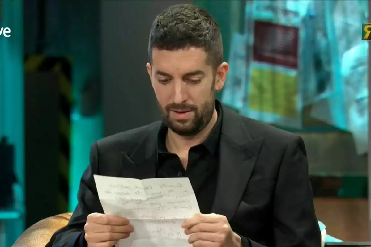 David Broncano con barba y cabello corto está leyendo una hoja de papel mientras está sentado en un entorno con iluminación tenue y fondo desenfocado. David Broncano con barba y cabello corto está leyendo una hoja de papel mientras está sentado en un entorno con iluminación tenue y fondo desenfocado.