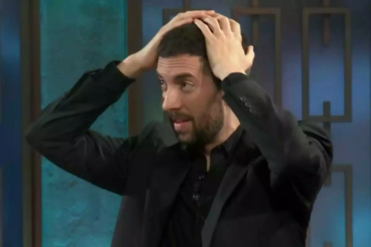 David Broncano con barba y cabello corto se toca la cabeza con ambas manos mientras mira hacia un lado en un entorno interior en La Revuelta.