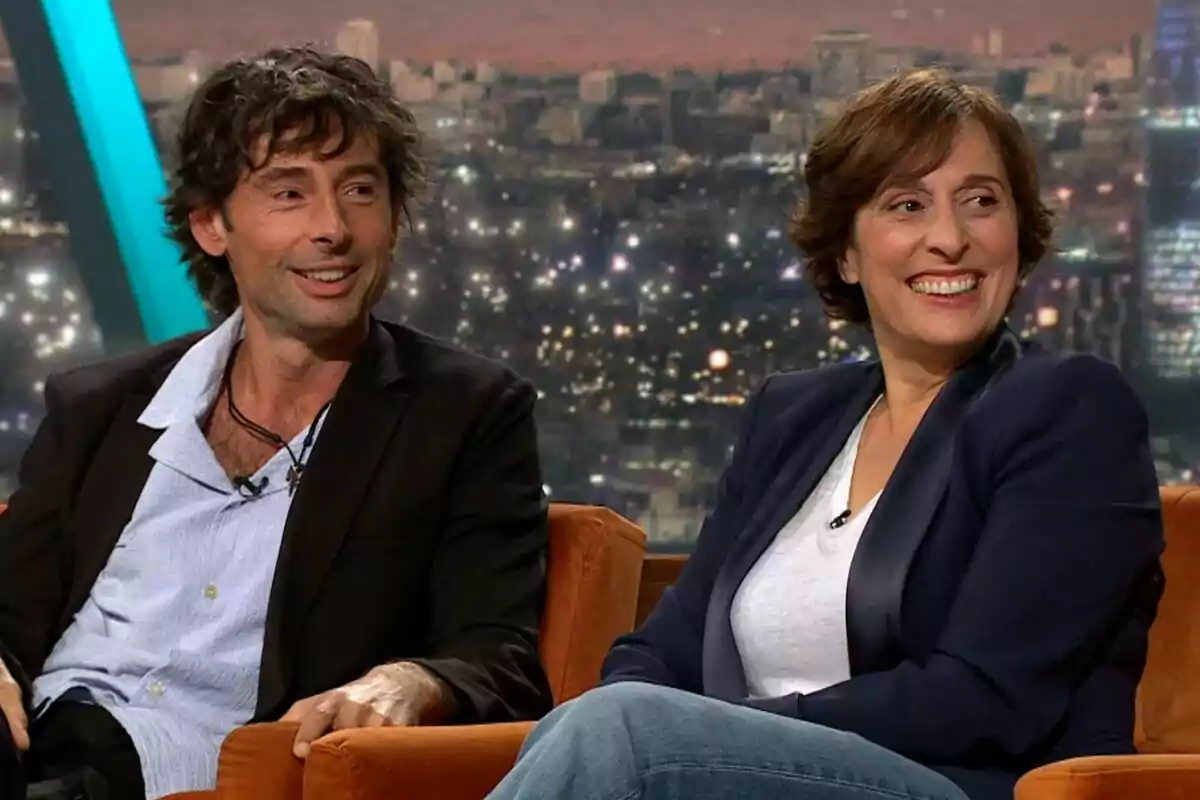 Bruno Oro i Clara Segura assegudes en un sofà durant una entrevista en un programa de televisió, Col·lapse, amb una ciutat il·luminada de fons.
