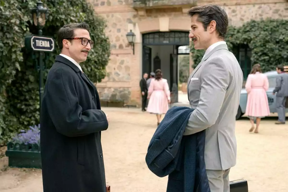 Bruno Tamarit y Alejandro Albarracín elegantemente vestidos conversan al aire libre frente a un edificio antiguo mientras varias personas caminan en el fondo en Sueños de Libertad