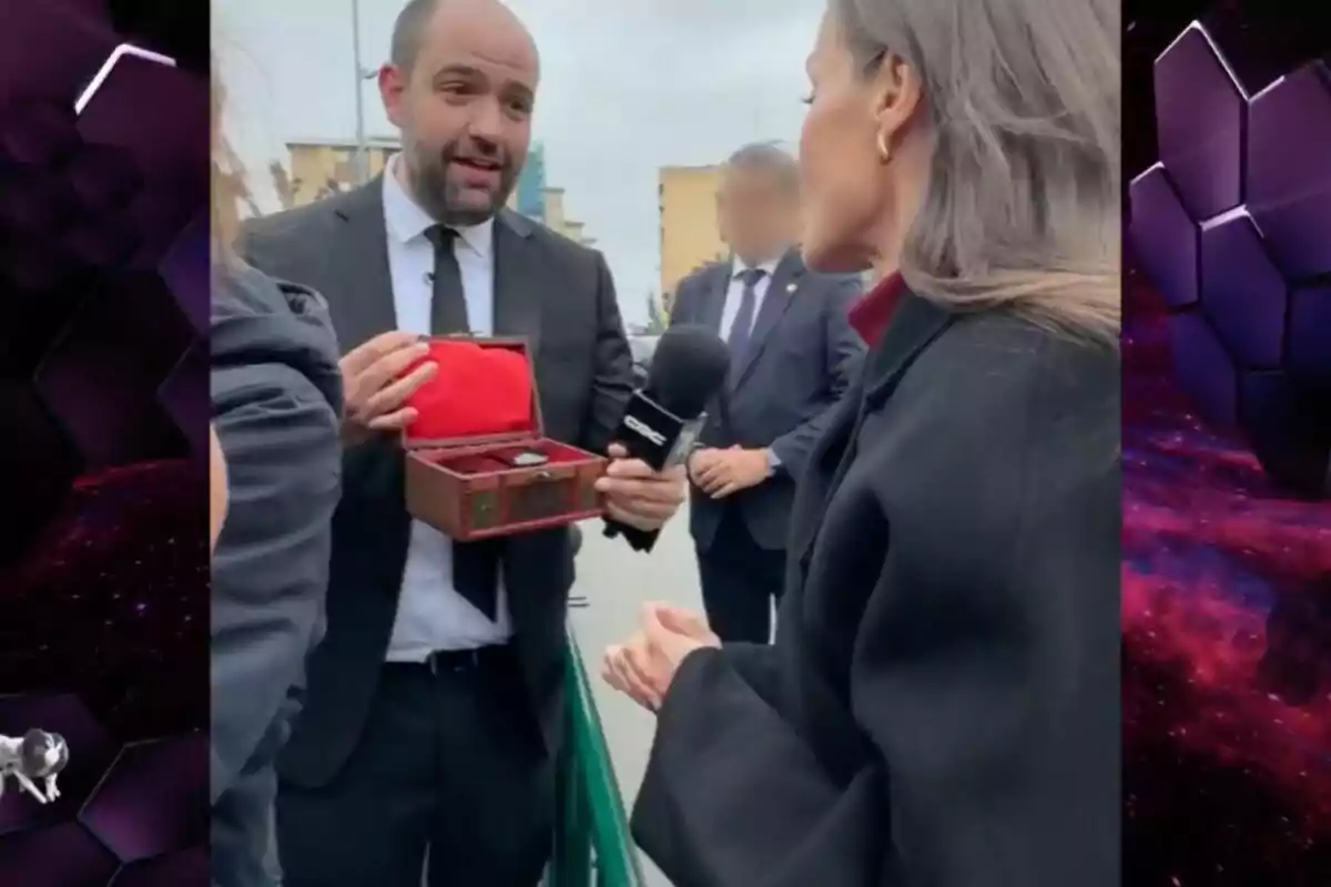 Daniel Fez amb vestit sosté una caixa oberta mentre parla amb la reina Letícia que porta un micròfon. Daniel Fez amb vestit sosté una caixa oberta mentre parla amb la reina Letícia que porta un micròfon.