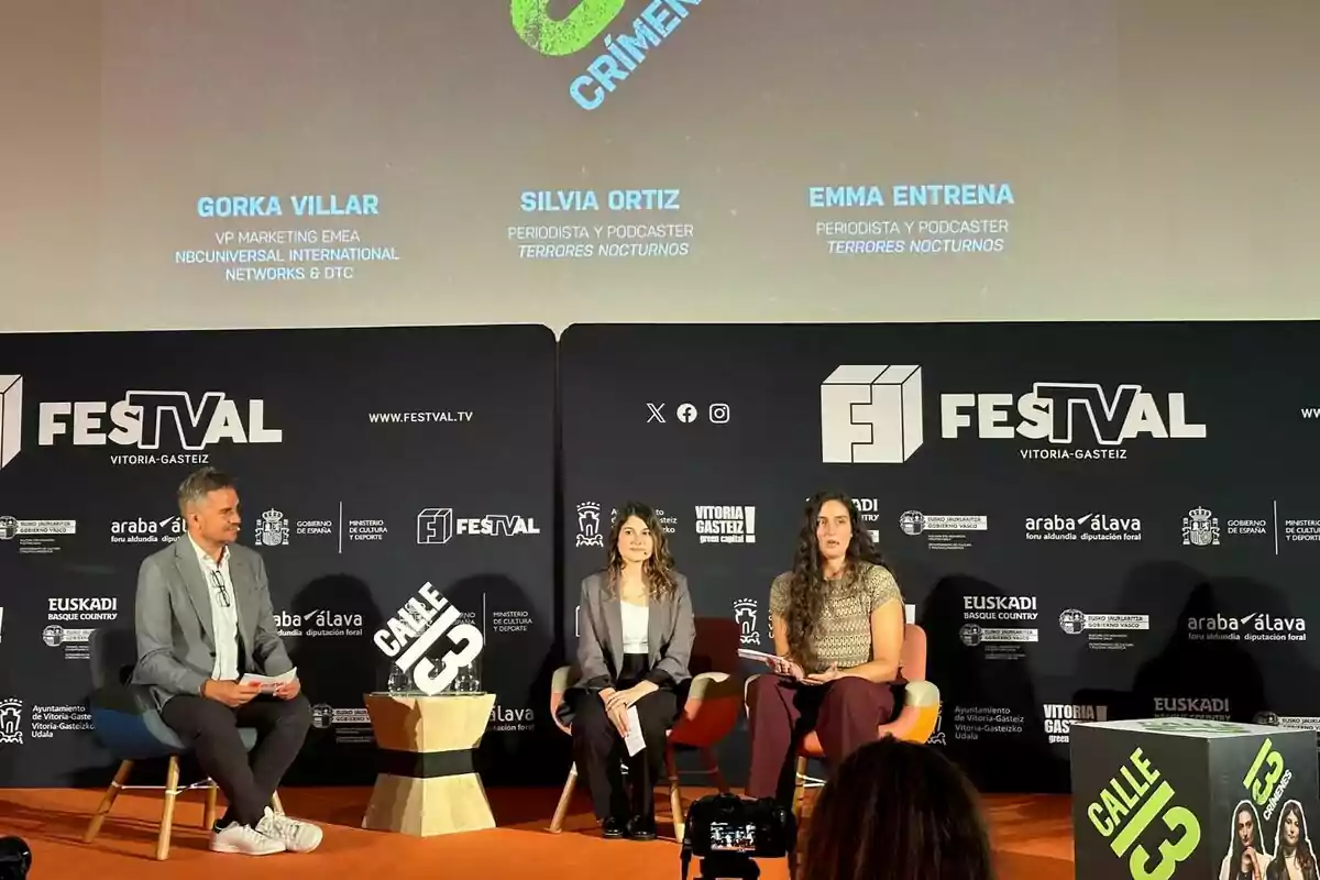 Tres personas están sentadas en un escenario durante un evento del FesTVal en Vitoria-Gasteiz, con un fondo que muestra los nombres Gorka Villar, Silvia Ortiz y Emma Entrena, junto con sus respectivos títulos y roles. Tres personas están sentadas en un escenario durante un evento del FesTVal en Vitoria-Gasteiz, con un fondo que muestra los nombres Gorka Villar, Silvia Ortiz y Emma Entrena, junto con sus respectivos títulos y roles.