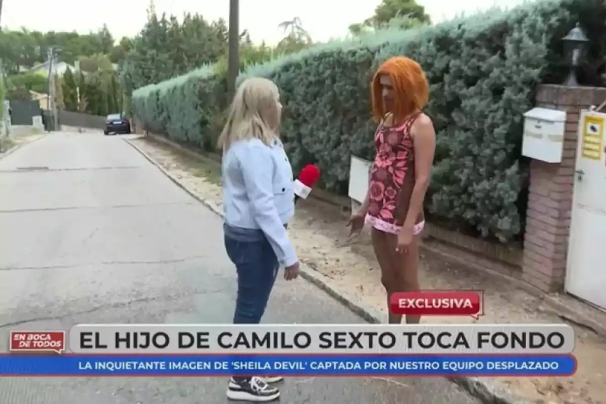 Una reportera de En Boca de Todos entrevista a Camilo Blanes con peluca naranja y vestido estampado en una calle residencial, con un seto y buzones al fondo. Una reportera de En Boca de Todos entrevista a Camilo Blanes con peluca naranja y vestido estampado en una calle residencial, con un seto y buzones al fondo.