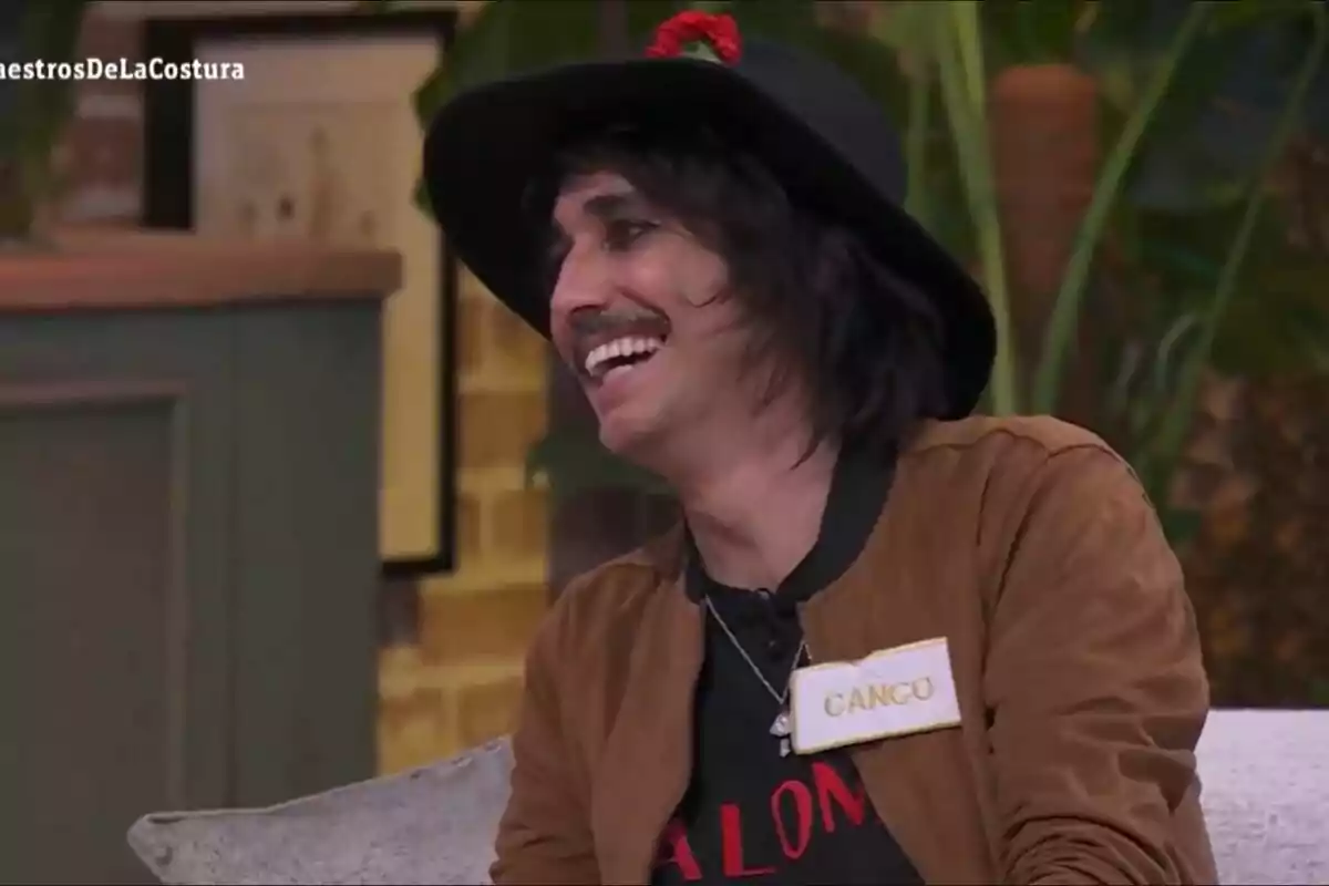 Canco Rodríguez sonriente con sombrero negro y chaqueta marrón, sentado en un ambiente interior en Maestros de la Costura Celebrity. Canco Rodríguez sonriente con sombrero negro y chaqueta marrón, sentado en un ambiente interior en Maestros de la Costura Celebrity.