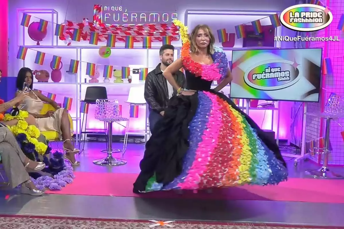 Un programa de televisión con temática de orgullo LGBT, donde una persona está modelando un vestido colorido con los colores del arcoíris, mientras otras personas observan y el set está decorado con banderas del orgullo. Un programa de televisión con temática de orgullo LGBT, donde una persona está modelando un vestido colorido con los colores del arcoíris, mientras otras personas observan y el set está decorado con banderas del orgullo.