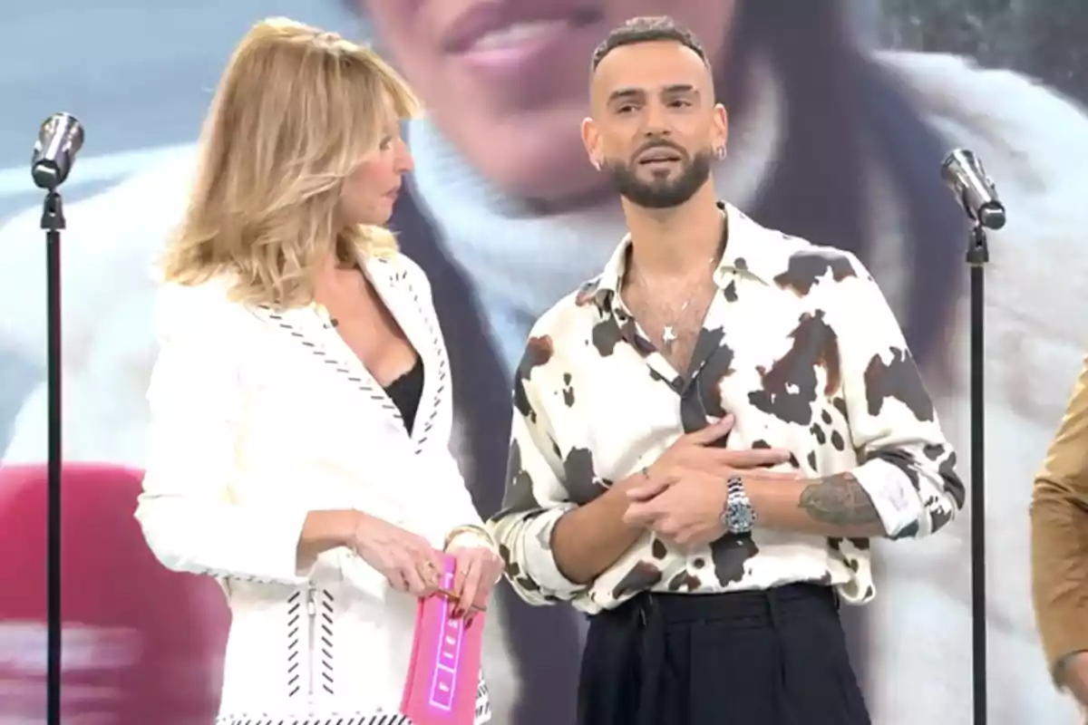 Manuel Cortés Bollo y Emma García de pie en un escenario con micrófonos, una mujer con traje blanco y un hombre con camisa estampada. Manuel Cortés Bollo y Emma García de pie en un escenario con micrófonos, una mujer con traje blanco y un hombre con camisa estampada.