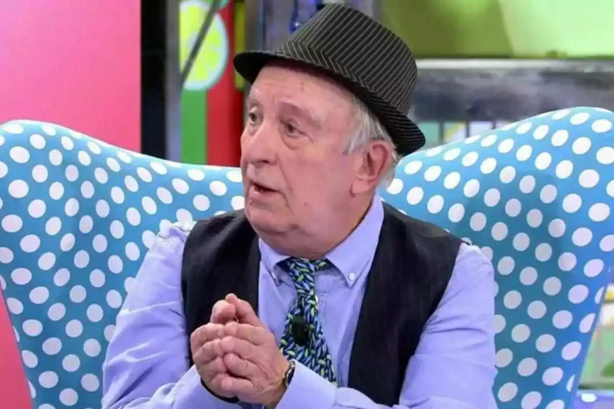 Carlos Ferrando amb barret de ratlles i armilla negra, assegut en una cadira amb disseny de pigues blanques sobre fons blau, parlant a Sálvame Carlos Ferrando amb barret de ratlles i armilla negra, assegut en una cadira amb disseny de pigues blanques sobre fons blau, parlant a Sálvame