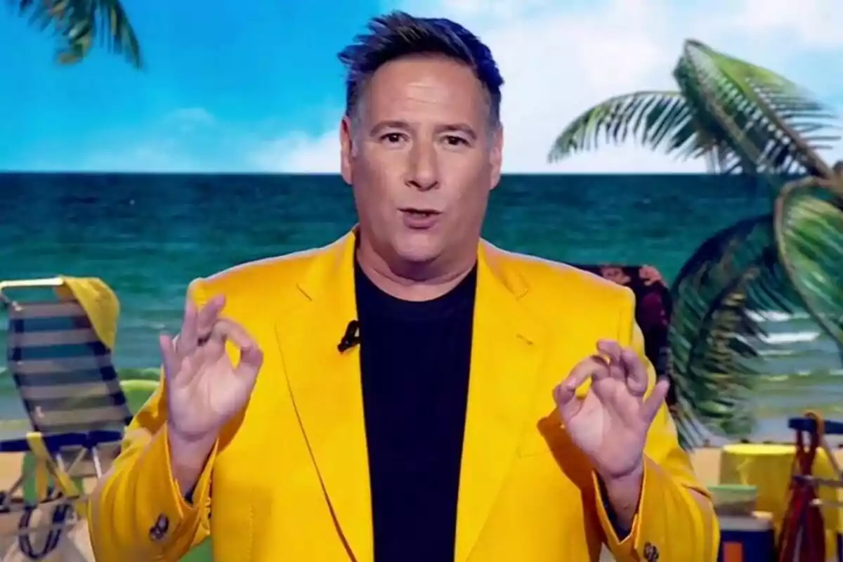 Carlos Latre amb jaqueta groga gesticulant amb les mans davant d'un fons de platja amb palmeres i una cadira de platja a Babylon Show Carlos Latre amb jaqueta groga gesticulant amb les mans davant d'un fons de platja amb palmeres i una cadira de platja a Babylon Show