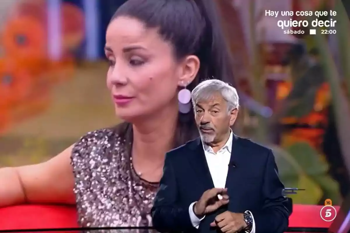Carlos Sobera de cabell canós i barba està parlant davant d'una pantalla que mostra Ana Herminia amb els cabells recollits i un vestit brillant a GH DÚO.