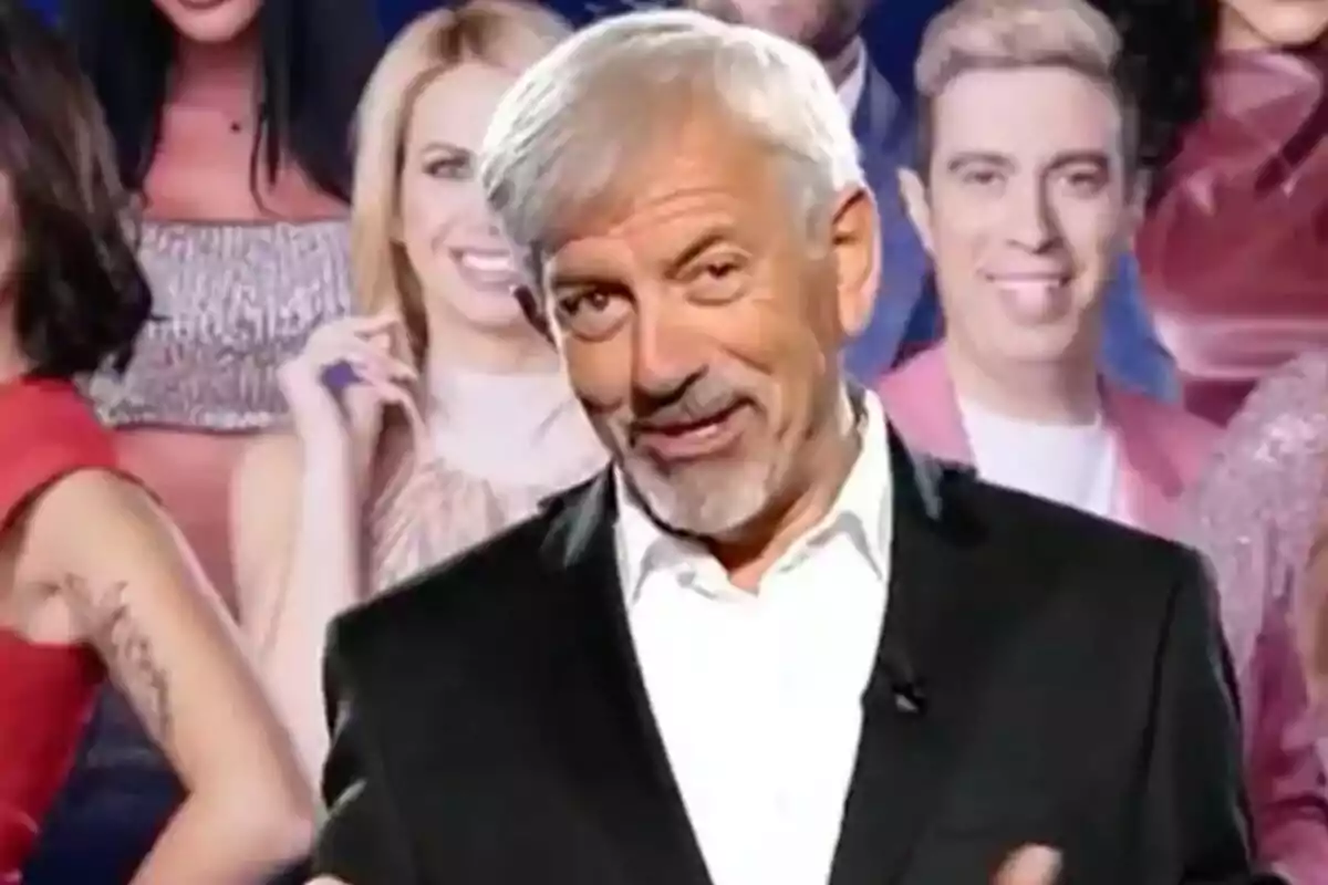 Carlos Sobera de cabello canoso y barba sonríe mientras lleva un traje oscuro y una camisa blanca, con un fondo de personas en ropa elegante en GH DÚO.