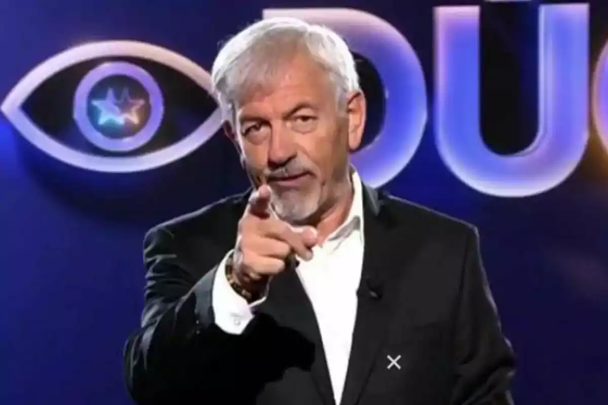 Carlos Sobera de cabello canoso y barba está señalando hacia la cámara con un fondo que tiene un logotipo de ojo y letras grandes de GH DÚO.