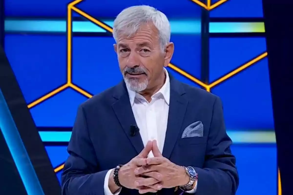 Carlos Sobera de cabello canoso y barba está de pie, vestido con un traje azul y camisa blanca, con un fondo azul y amarillo geométrico en el plató de Todos por ti. Carlos Sobera de cabello canoso y barba está de pie, vestido con un traje azul y camisa blanca, con un fondo azul y amarillo geométrico en el plató de Todos por ti.