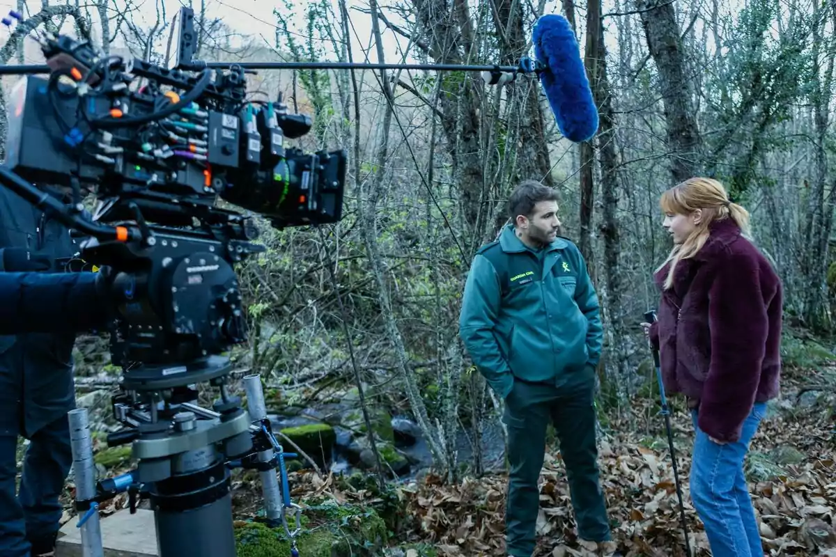 Una escena de rodaje en un bosque donde Alfons Nieto con uniforme verde y Catalina Sopelana con chaqueta marrón están conversando mientras una cámara de cine y un micrófono capturan el momento en El cuco de cristal.