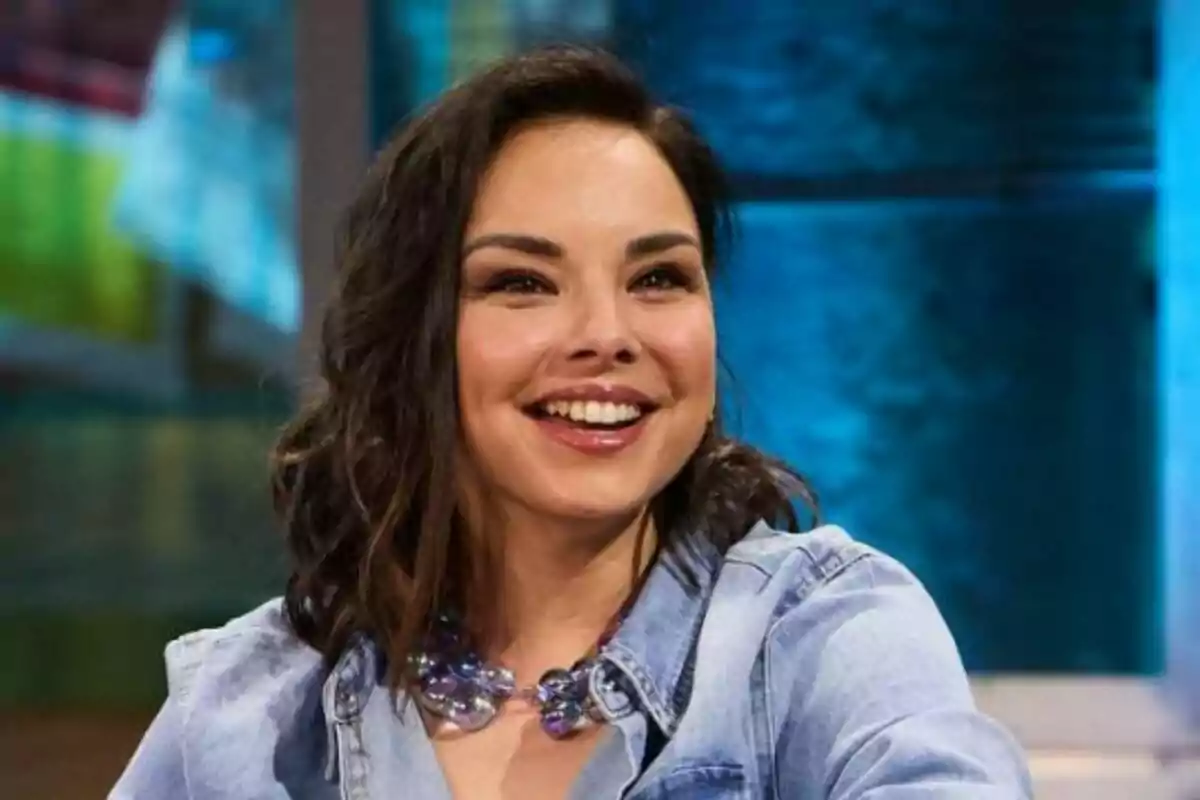 Chenoa sonriendo con una camisa de mezclilla y un collar de cuentas en un entorno interior con fondo azul en La Revuelta.