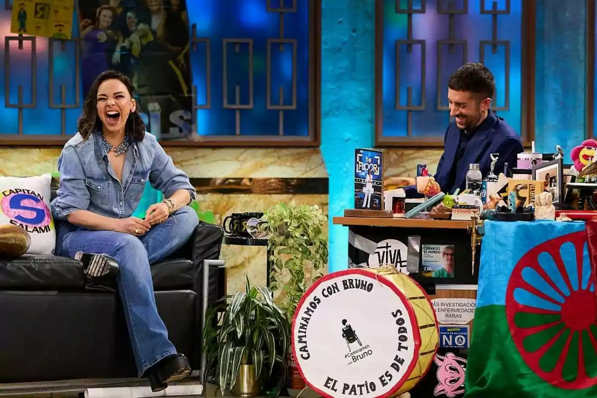Chenoa riéndose sentada en un sofá junto a David Broncano en una mesa llena de objetos en La Revuelta con un fondo azul y marrón. Chenoa riéndose sentada en un sofá junto a David Broncano en una mesa llena de objetos en La Revuelta con un fondo azul y marrón.