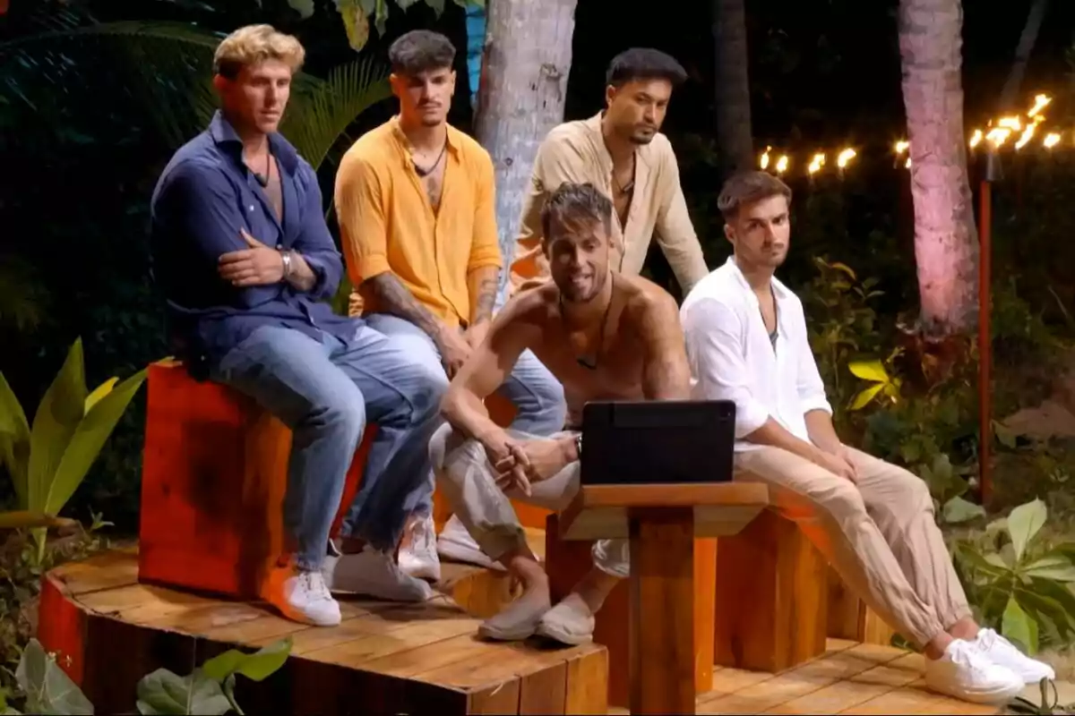 Cinco hombres sentados, Montoya sin camiseta, en un entorno al aire libre, rodeados de vegetación y luces, observan una pantalla con atención La Isla de las Tentaciones. Cinco hombres sentados, Montoya sin camiseta, en un entorno al aire libre, rodeados de vegetación y luces, observan una pantalla con atención La Isla de las Tentaciones.