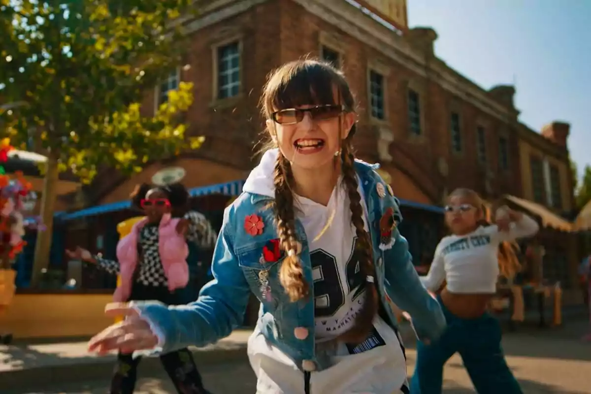 Chloe DeLaRosa con gafas y trenzas sonríe mientras baila en una calle con un edificio de ladrillo al fondo, acompañada de otras personas que también están bailando. Chloe DeLaRosa con gafas y trenzas sonríe mientras baila en una calle con un edificio de ladrillo al fondo, acompañada de otras personas que también están bailando.