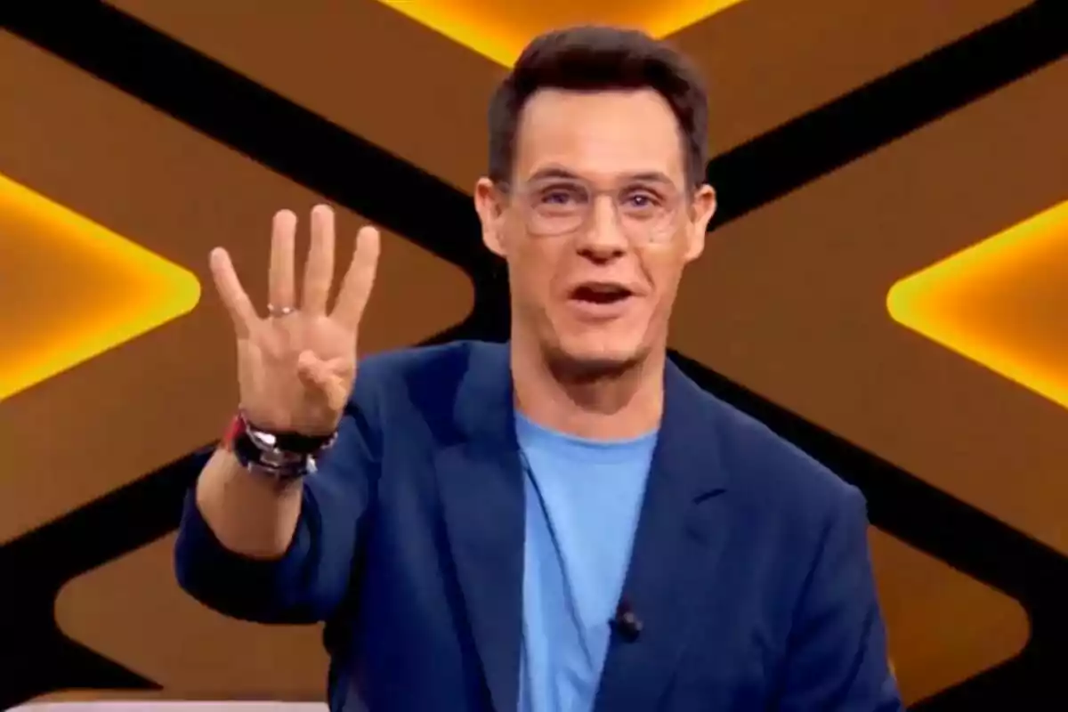 Christian Gálvez amb ulleres i jaqueta blava aixeca quatre dits davant d'un fons amb llums grogues a Boom. Christian Gálvez amb ulleres i jaqueta blava aixeca quatre dits davant d'un fons amb llums grogues a Boom.