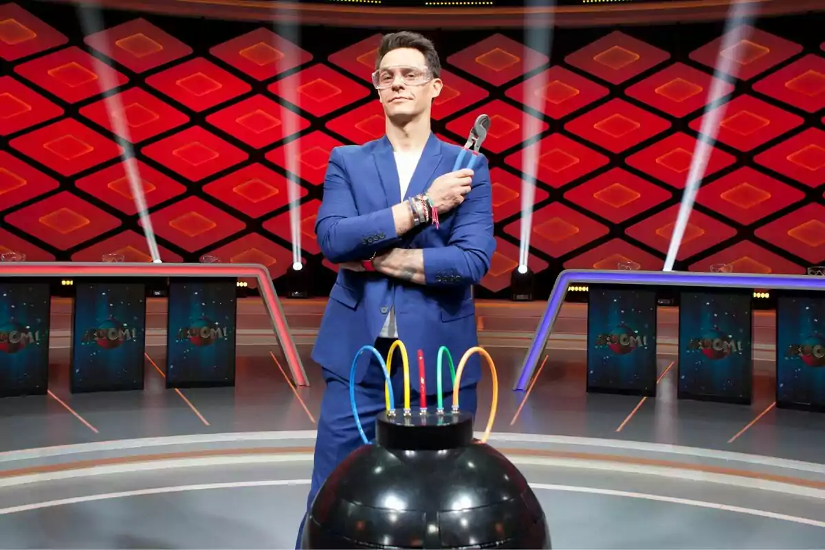 Christian Gálvez amb ulleres de seguretat i vestit blau sosté un parell d'alicates al plató de Boom! amb un fons vermell i negre, llums brillants i una taula amb cables de colors. Christian Gálvez amb ulleres de seguretat i vestit blau sosté un parell d'alicates al plató de Boom! amb un fons vermell i negre, llums brillants i una taula amb cables de colors.