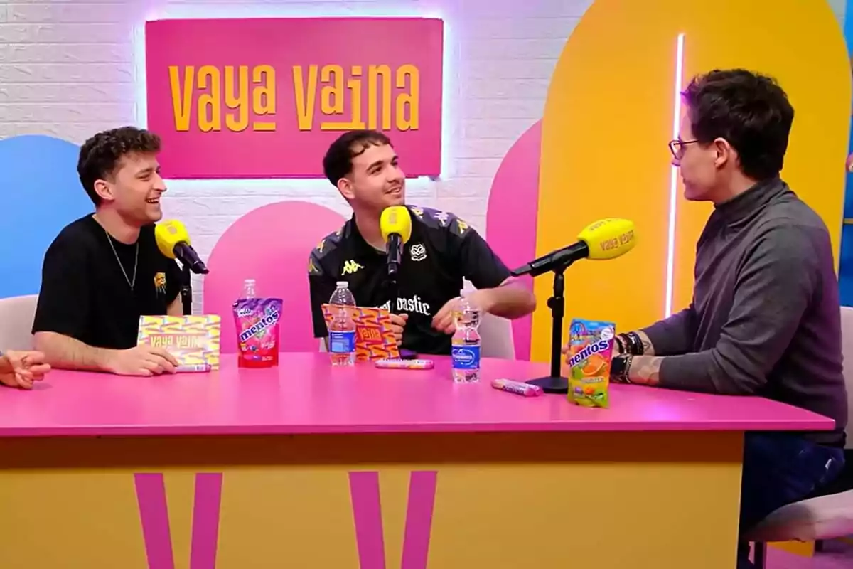 Christian Gálvez, Alfon de la Rocha y Dani Gordo conversan animadamente en un set de grabación colorido con micrófonos amarillos y productos sobre la mesa, bajo un letrero que dice Christian Gálvez, Alfon de la Rocha y Dani Gordo conversan animadamente en un set de grabación colorido con micrófonos amarillos y productos sobre la mesa, bajo un letrero que dice
