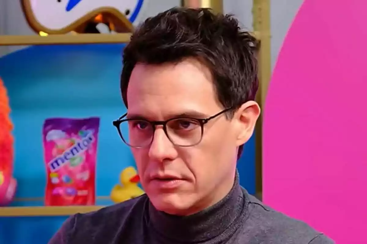 Christian Gálvez con gafas y suéter gris oscuro en una entrevista en Vaya Vaina está en un entorno colorido con un paquete de Mentos y un pato de goma en el fondo.