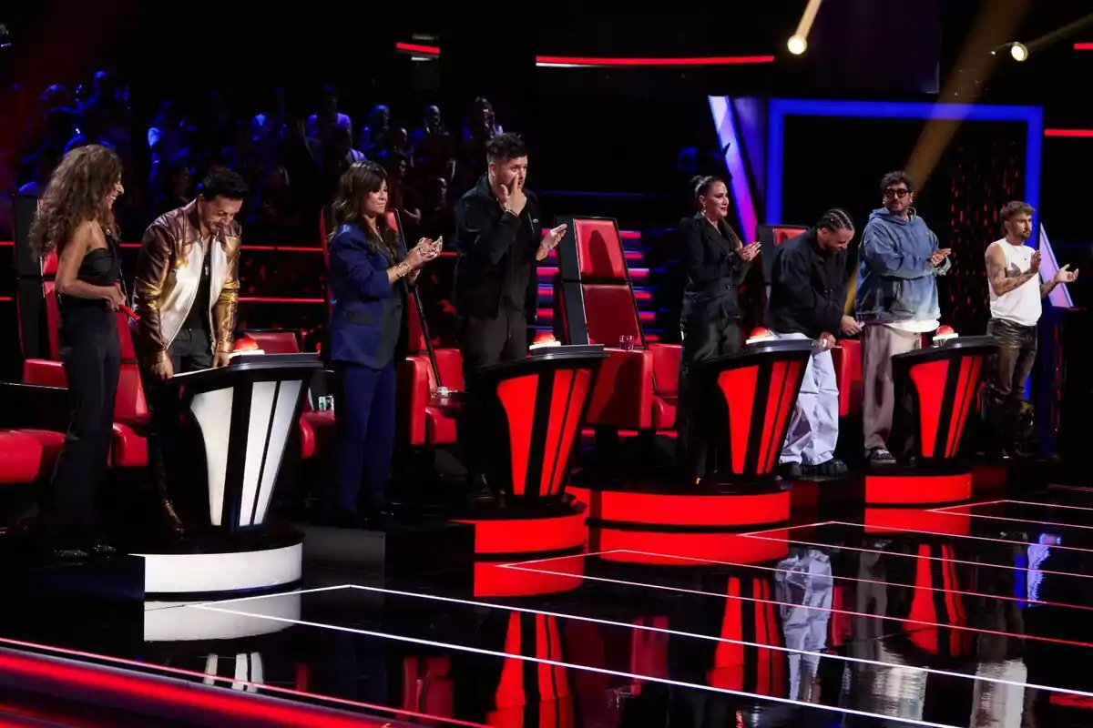 Els coaches i assessors de La Voz drets al costat de cadires vermelles en un escenari il·luminat amb llums brillants i una audiència al fons: Rosario Flores, Lluís Fonsi, Vanesa Martín, Pablo López, Malú, Prince Royce, Antonio Orozco i Dani Fernández.