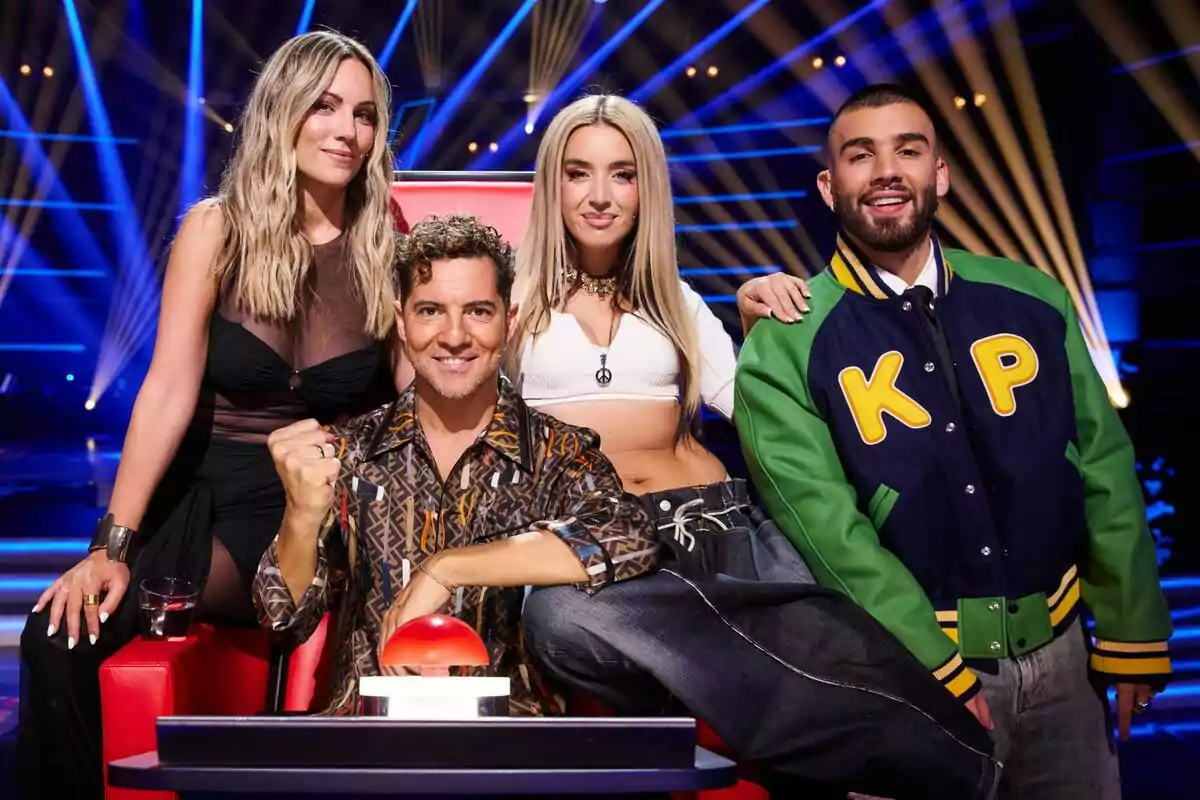 Edurne, David Bisbal, Lola Índigo i Manuel Turizo posen junts en un escenari il·luminat amb llums blaves i grogues, una d'elles està asseguda en una cadira vermella amb un botó gran davant d'ells a La Voz Kids.