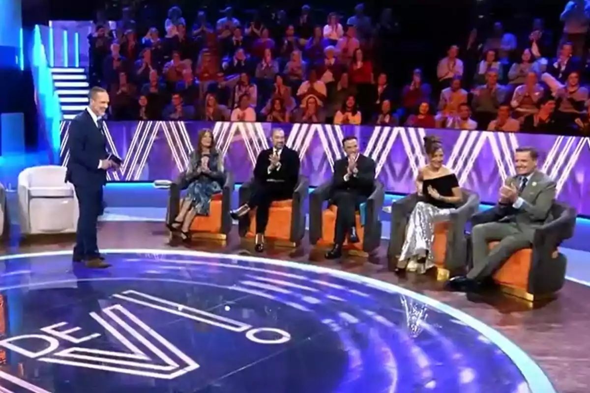 Un grupo de personas está sentado en De Viernes, Santi Acosta de pie frente a Ángela Portero, José Antonio León, Antonio Rossi, Patricia Pérez y Antonio Montero, mientras el público aplaude desde las gradas. Un grupo de personas está sentado en De Viernes, Santi Acosta de pie frente a Ángela Portero, José Antonio León, Antonio Rossi, Patricia Pérez y Antonio Montero, mientras el público aplaude desde las gradas.