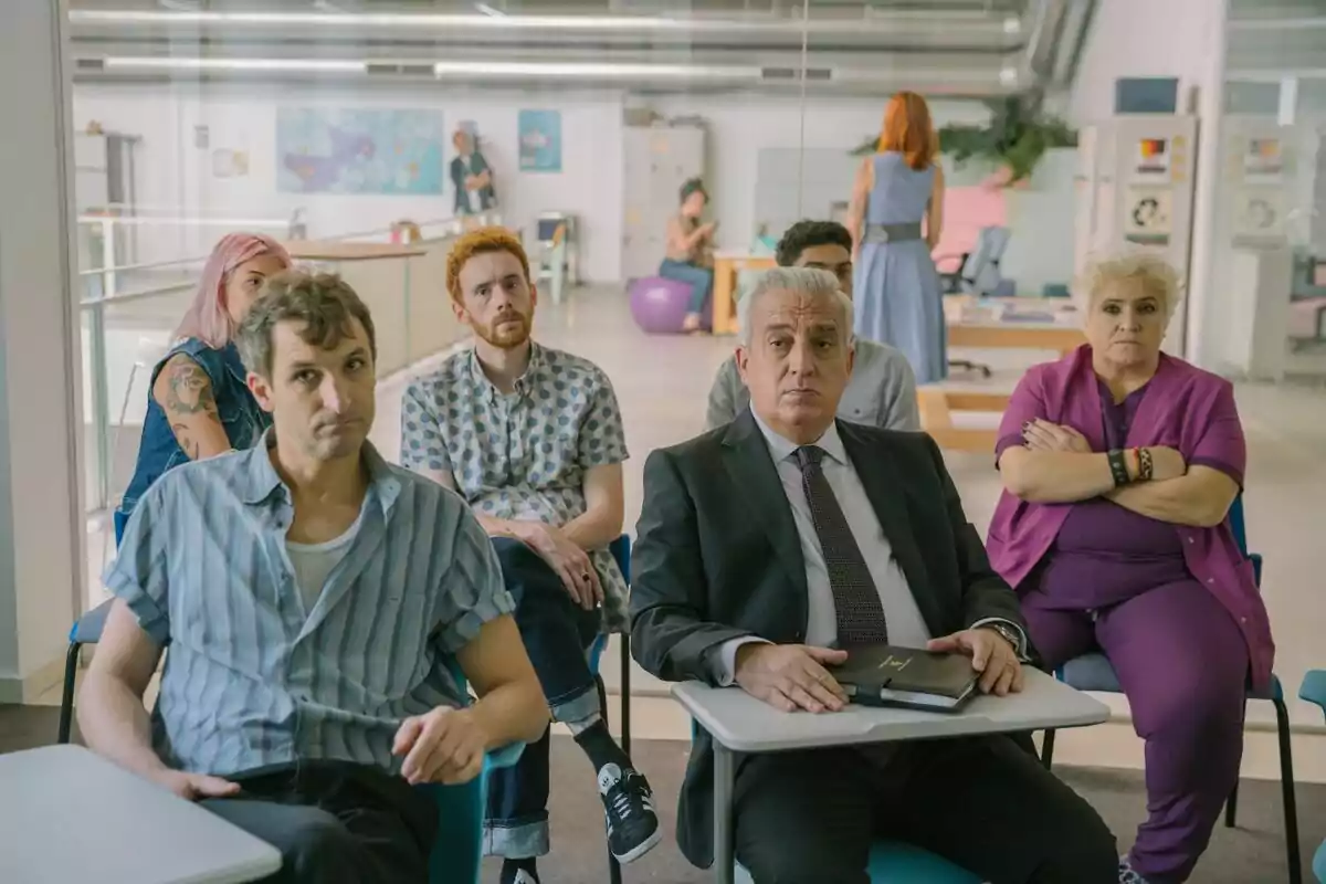 Un grupo de personas encabezado por Leo Harlem está sentado en un aula, algunas parecen atentas mientras otras tienen expresiones serias o pensativas, en un entorno que parece ser una oficina moderna.