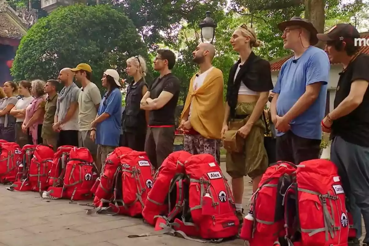 Un grup de concursants de peu en fila al costat de motxilles vermelles en un entorn a l'aire lliure amb arbres i un edifici al fons a la promo de Pekin Express. Un grup de concursants de peu en fila al costat de motxilles vermelles en un entorn a l'aire lliure amb arbres i un edifici al fons a la promo de Pekin Express.