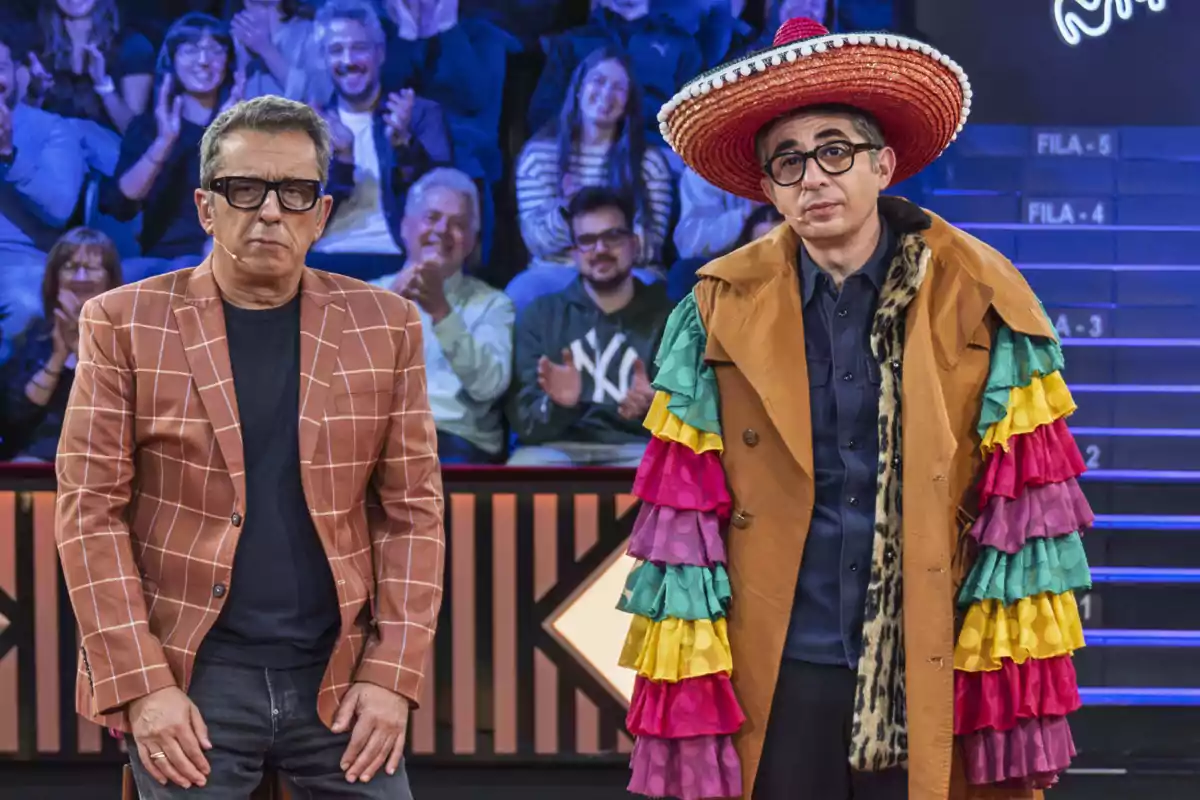 Andreu Buenafuente i Berto Romero estan drets en un escenari, un porta un barret gran i un abric amb volants de colors, mentre que l'altre porta una jaqueta de quadres; al fons, el públic aplaudeix a El Consultori de Berto. Andreu Buenafuente i Berto Romero estan drets en un escenari, un porta un barret gran i un abric amb volants de colors, mentre que l'altre porta una jaqueta de quadres; al fons, el públic aplaudeix a El Consultori de Berto.