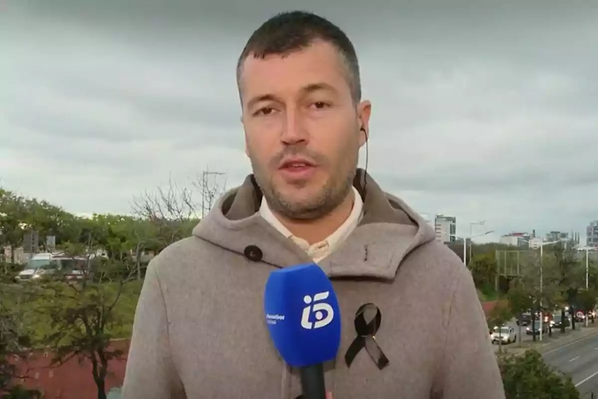 Un reportero de Informativos Telecinco con un micrófono azul y un lazo negro en su abrigo está de pie al aire libre con una carretera y árboles al fondo. Un reportero de Informativos Telecinco con un micrófono azul y un lazo negro en su abrigo está de pie al aire libre con una carretera y árboles al fondo.