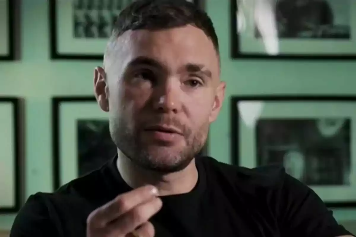 Cristian Suescun con barba corta y cabello oscuro está hablando mientras gesticula con la mano, con un fondo de cuadros en la pared. Cristian Suescun con barba corta y cabello oscuro está hablando mientras gesticula con la mano, con un fondo de cuadros en la pared.