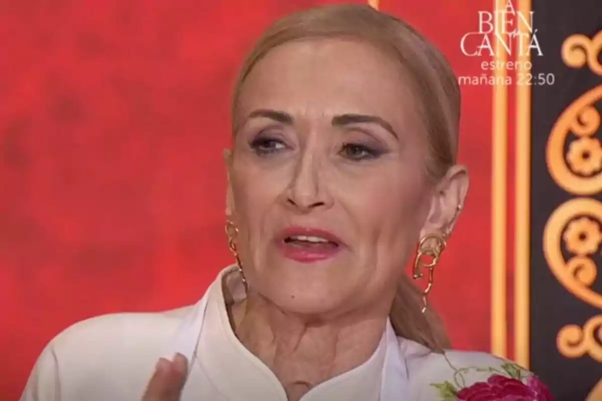 Cristina Cifuentes amb cabell ros recollit i pendents daurats, parlant davant d'un fons vermell amb detalls decoratius a MasterChef Celebrity. Cristina Cifuentes amb cabell ros recollit i pendents daurats, parlant davant d'un fons vermell amb detalls decoratius a MasterChef Celebrity.