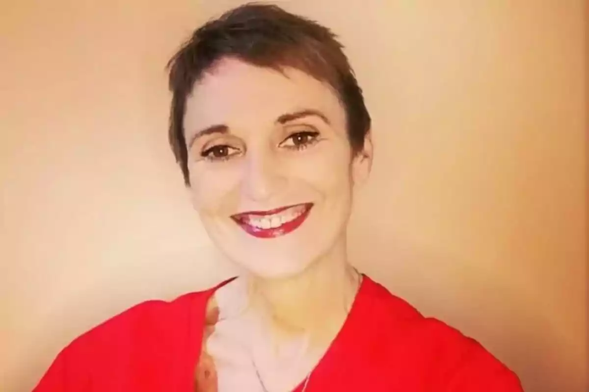 Cristina Fenollar, de cabell curt i fosc, somriu a la càmera, porta una samarreta vermella i es troba davant d’un fons beix.