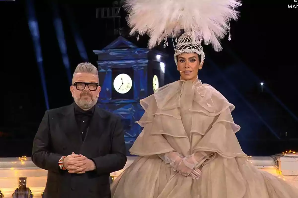Alberto Chicote y Cristina Pedroche elegantemente vestidas en un evento nocturno con un reloj iluminado de fondo en las Campanadas.