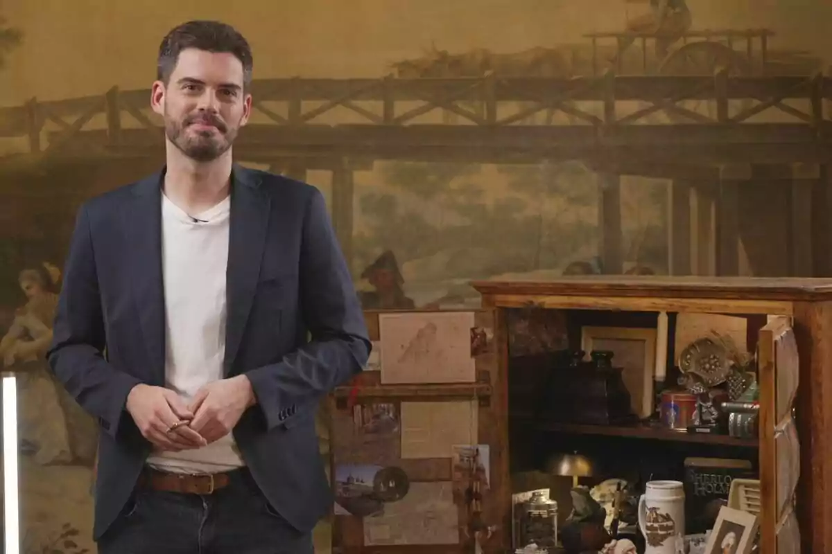 Fran Izuzquiza con barba y chaqueta oscura está de pie frente a un fondo que parece ser una pintura antigua, junto a un mueble de madera lleno de objetos decorativos y libros en Cuarto de Maravillas.